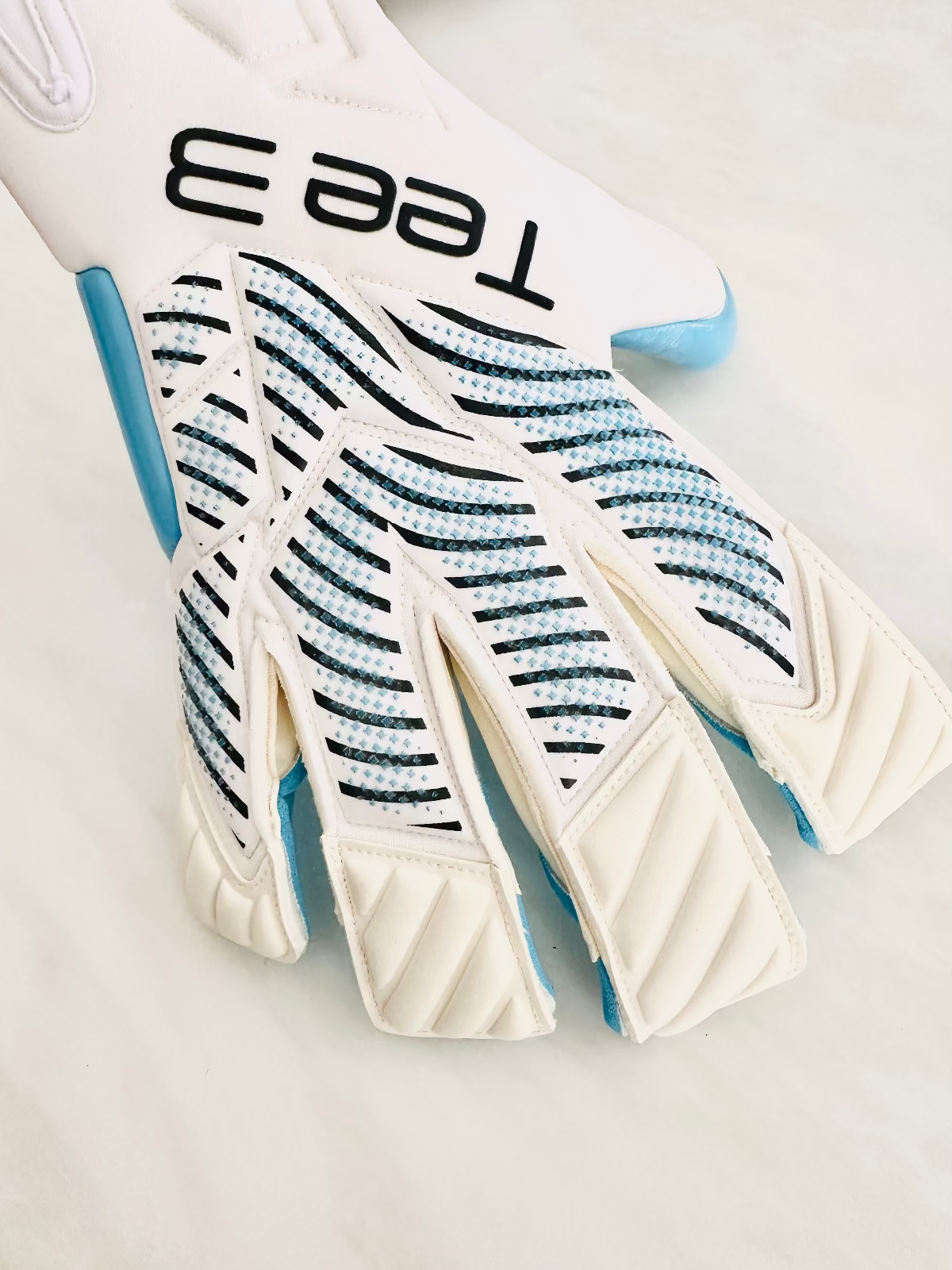 A close-up of a white goalkeeper glove with black 'Tee 3' branding, featuring blue and black striped knit finger protectors and light blue accents.
