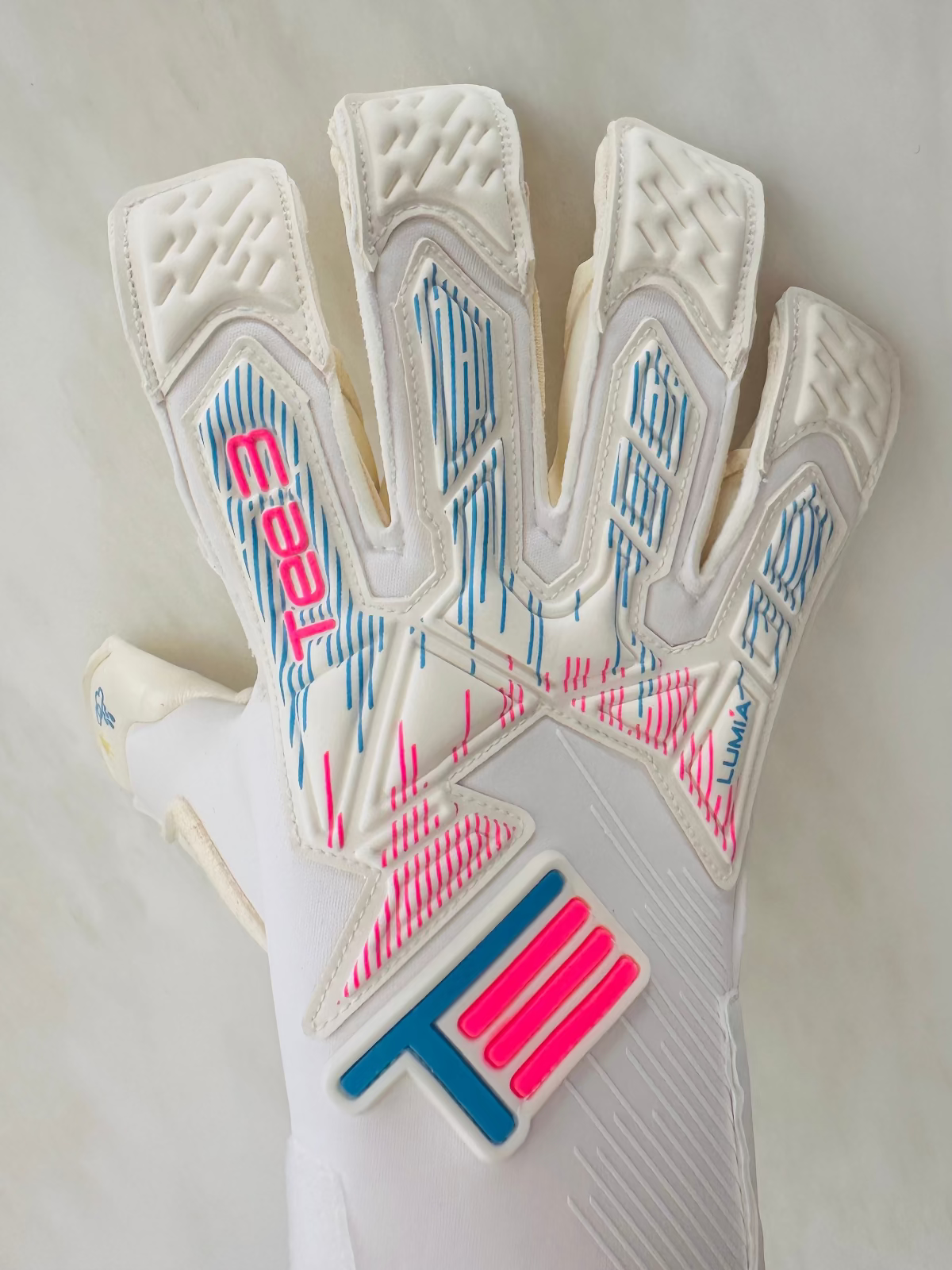 A close-up of a white Tee3 Lumia goalkeeper glove showing the back of the hand with textured finger panels, blue and pink graphic line details, and a large pink and blue 'T3' logo patch.