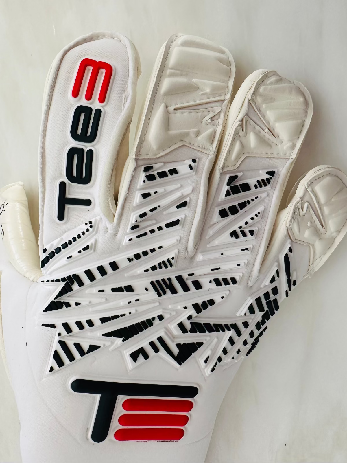 A close-up of a white TEE-3 Goalkeeping professional goalkeeper glove with red and black raised punch pads, made from neoprene with specific white and black accents and details. 