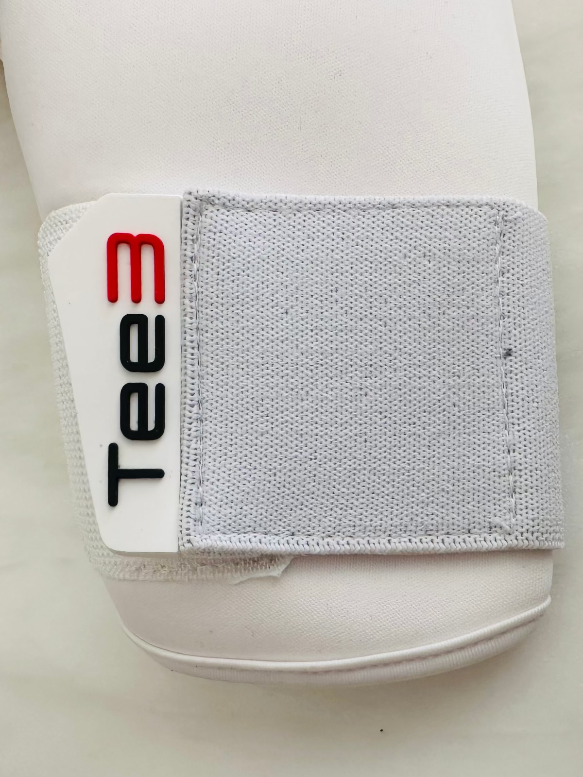 A close-up of a white TEE-3 Goalkeeping professional goalkeeper glove wrist strap with red and black raised Tee3 details on the pull tab and velcro to fasten 