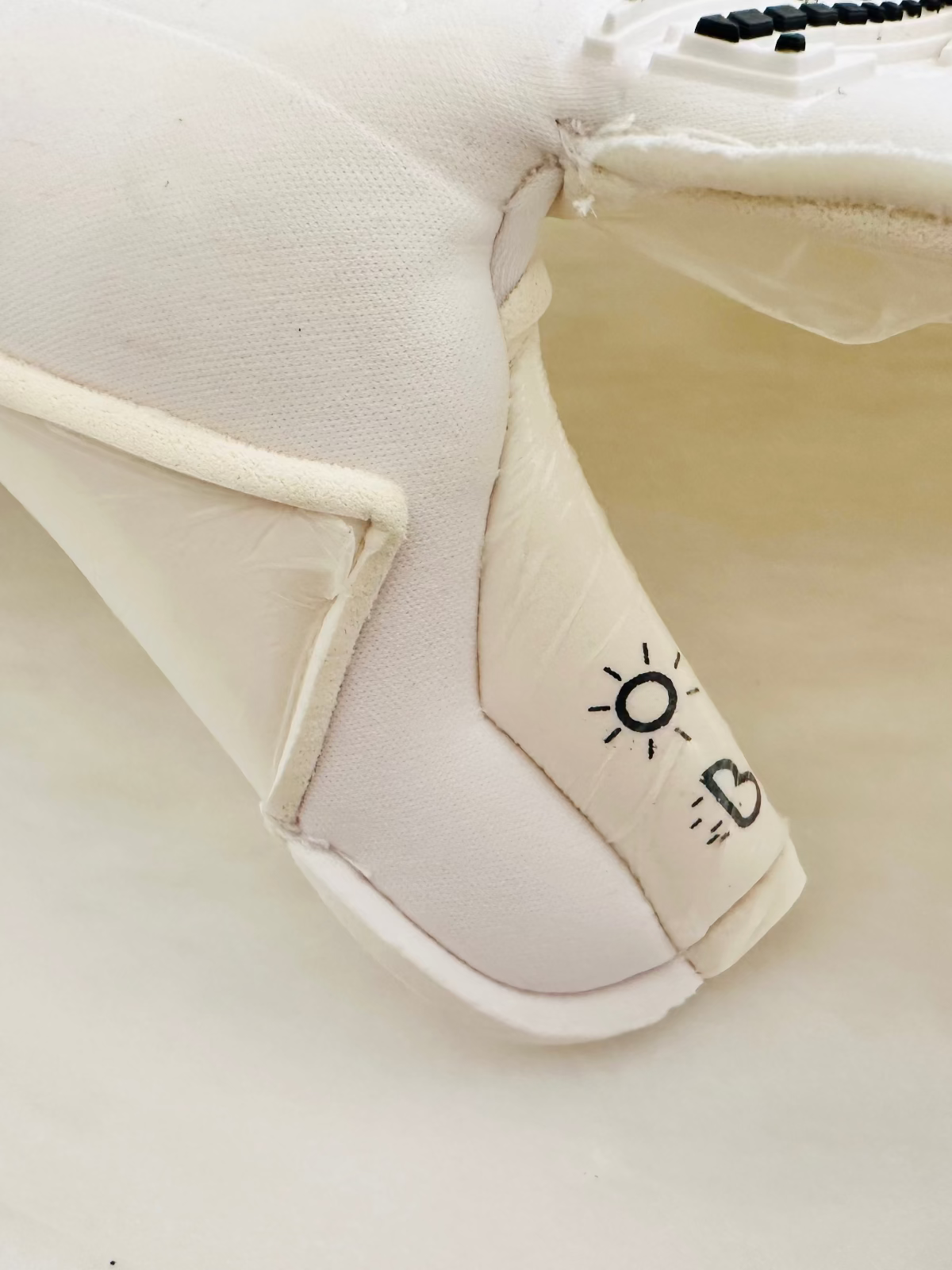 A close-up of a white TEE-3 professional goalkeeping brand double wrapped thumb with the rain and sun icons printed showing that it can be used in wet weather and warm weather made from neoprene