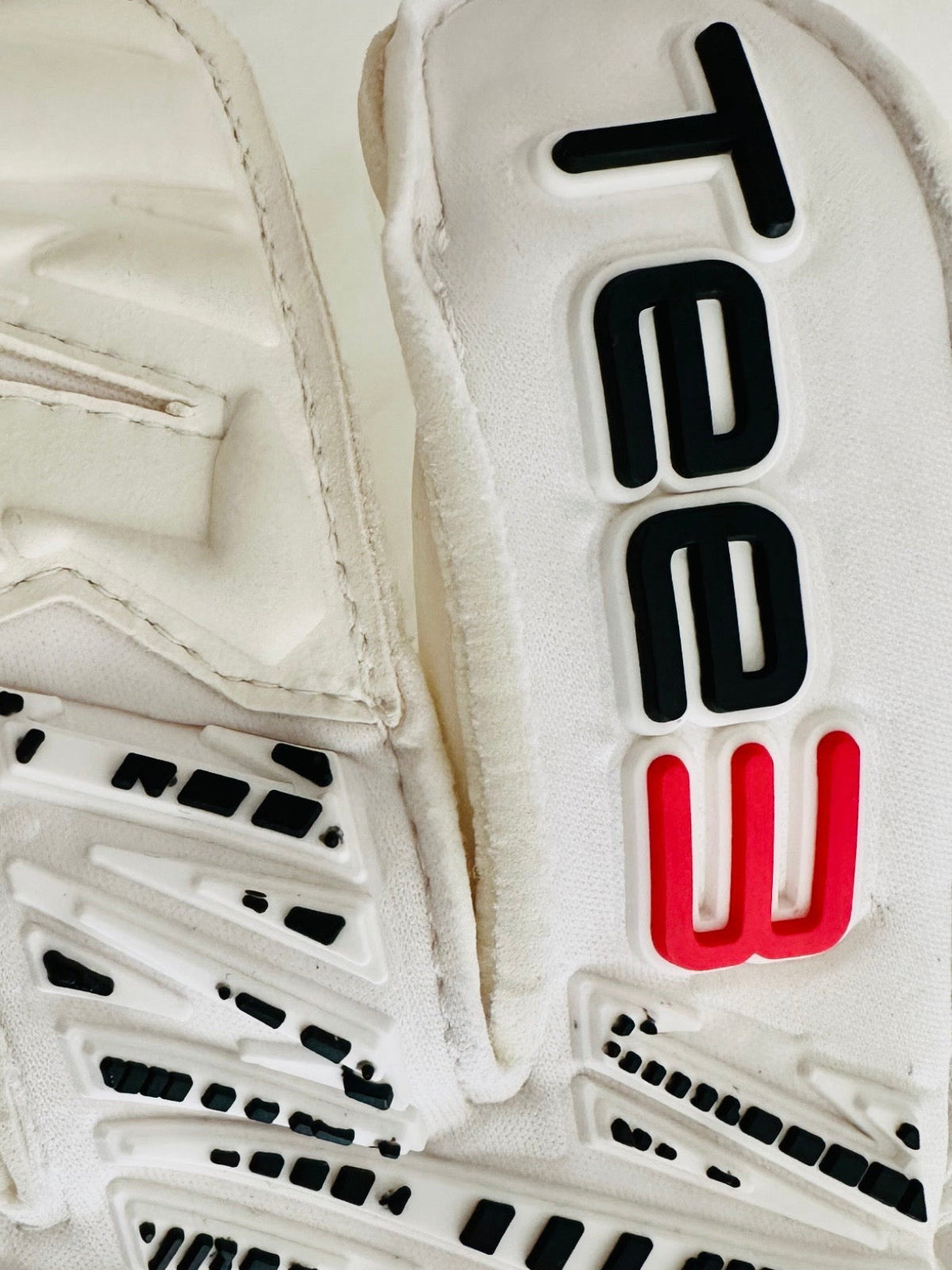 Close-up of a white athletic glove with black and red 'Tee3' branding on the back, showing textured white and black grip patterns on the palm side.