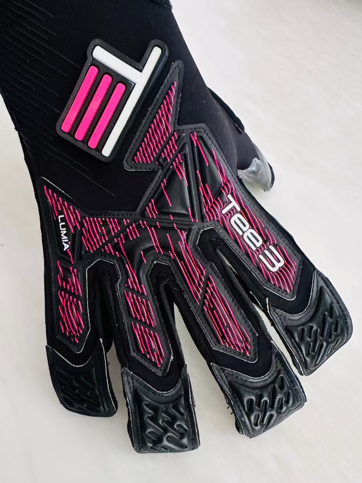 A black TEE-3 professional goalkeeping brand minimalist goalkeeper glove with pink and white graphic accents, black contact latex featuring the 'LUMIA' model name on the side, photographed on a light neutral background. 