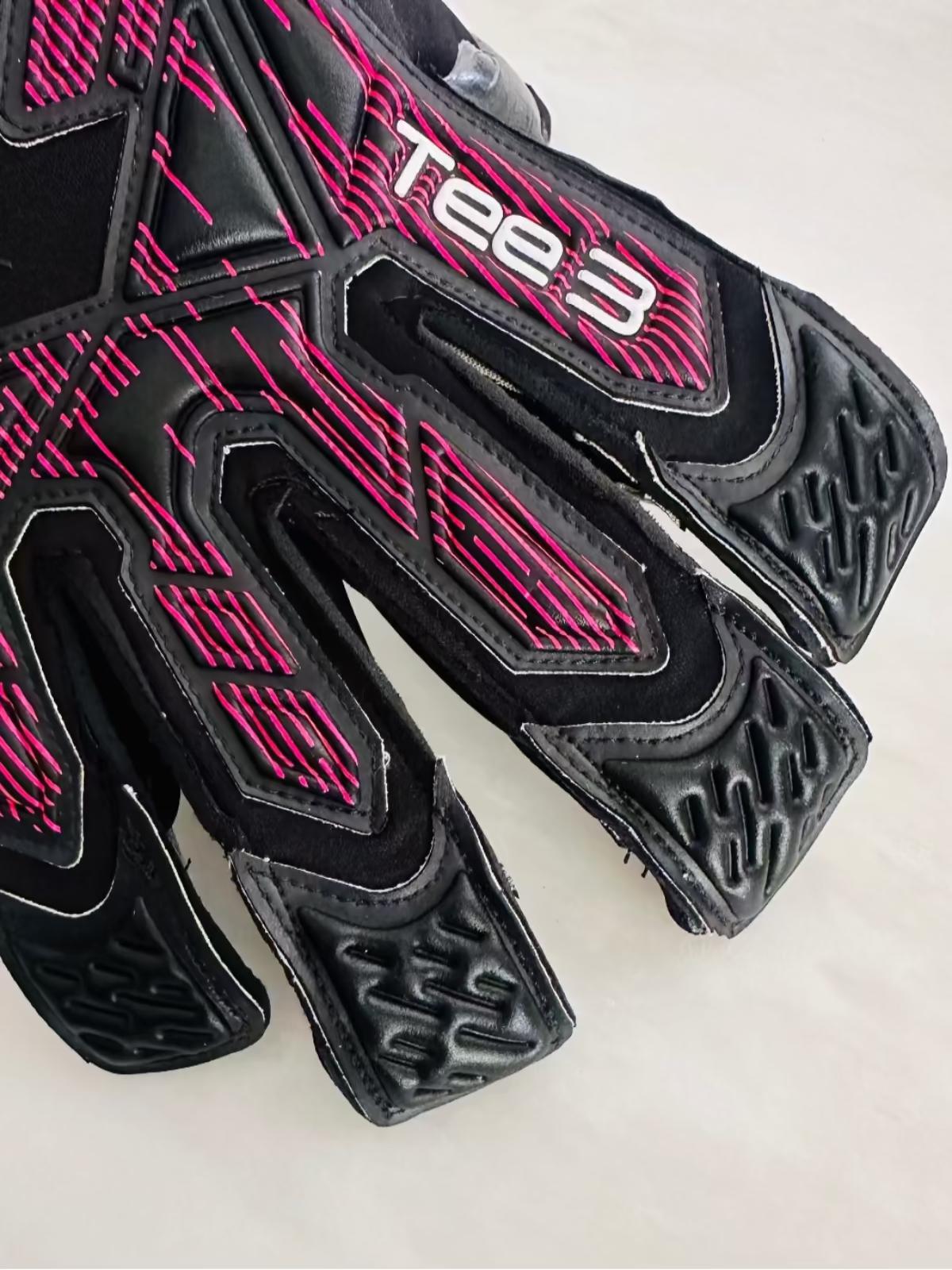 A close-up of a black TEE-3 professional goalkeeping brand minimalist goalkeeper glove with pink and white graphic accents, black contact latex black soccer goalkeeper glove with vibrant pink accents and the brand name 'Tee 3' printed on the side, resting on a light off-white surface.