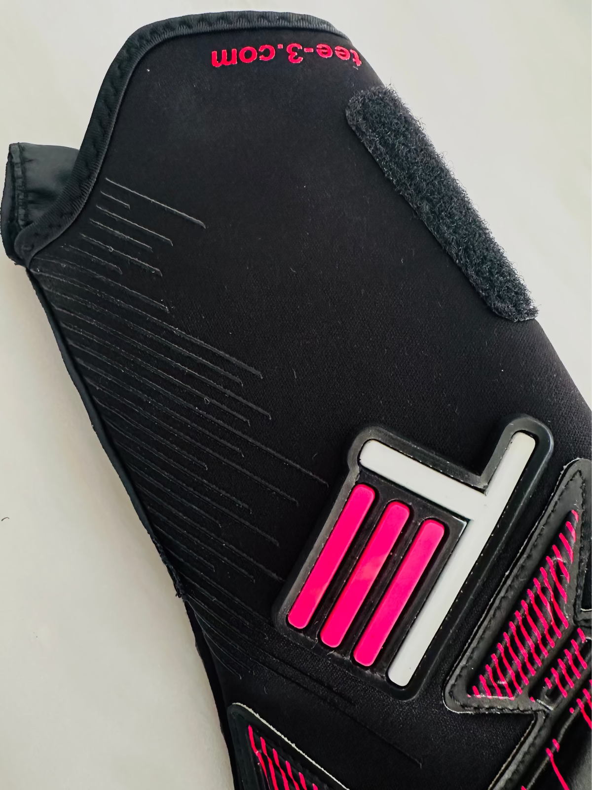 A close-up of a black TEE-3 professional goalkeeping brand wrist entry minimalist goalkeeper glove with pink and white graphic accents, made from neoprene with specific accents and details. The website address "www.tee-3.com" is printed at the beginning of the tunnel 