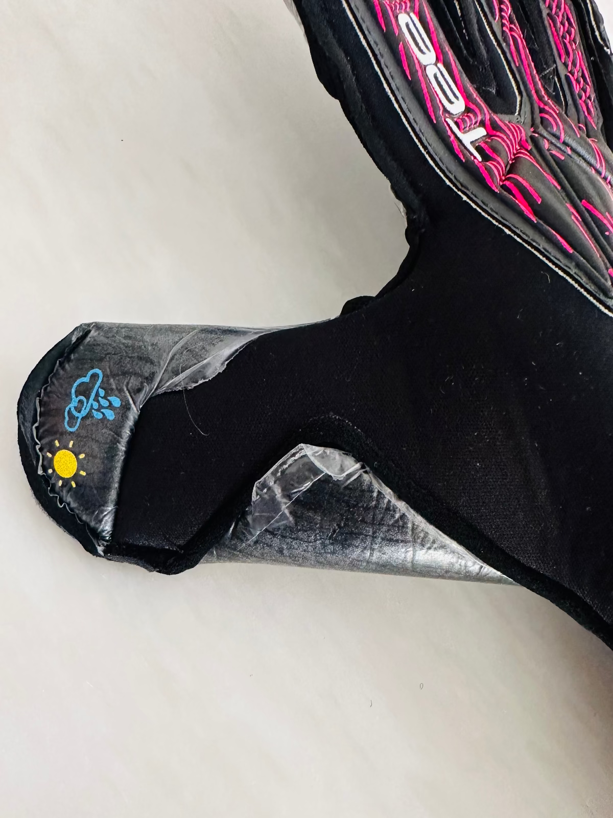 A close-up of a black TEE-3 professional goalkeeping brand double wrapped thumb with the rain and sun icons printed showing that it can be used in wet weather and warm weather 